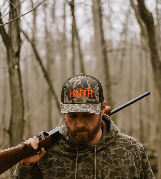 Hunter wearing a camouflage hoodie holding a rifle over their right shoulder. The hunter wears a hat that reads "HNTR".
