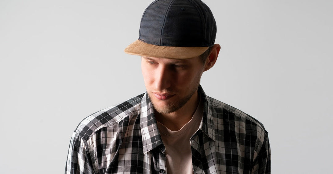 A young man wearing a black and white plaid shirt has a black snapback hat featuring a brown brim on his head.
